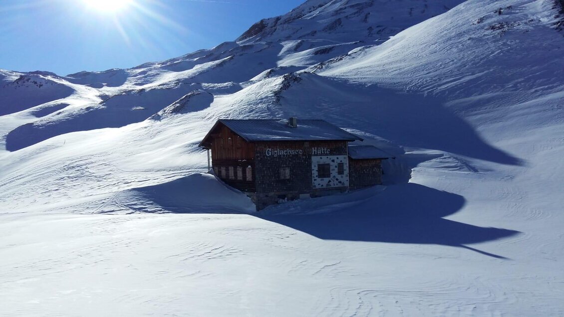 Eine gemütliche Hütte steht inmitten einer verschneiten Landschaft. Die Berge im Hintergrund strahlen unter dem klaren blauen Himmel. | © Giglachseehütte