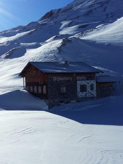 A cozy cabin stands in the middle of a snowy landscape. The mountains in the background shine under the clear blue sky. | © Giglachseehütte