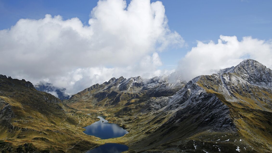 Eine beeindruckende Berglandschaft mit schneebedeckten Gipfeln und tiefen Tälern. Im Vordergrund liegt ein klarer See, umgeben von grünen Wiesen und dramatischen Wolken. | © Herbert Raffalt
