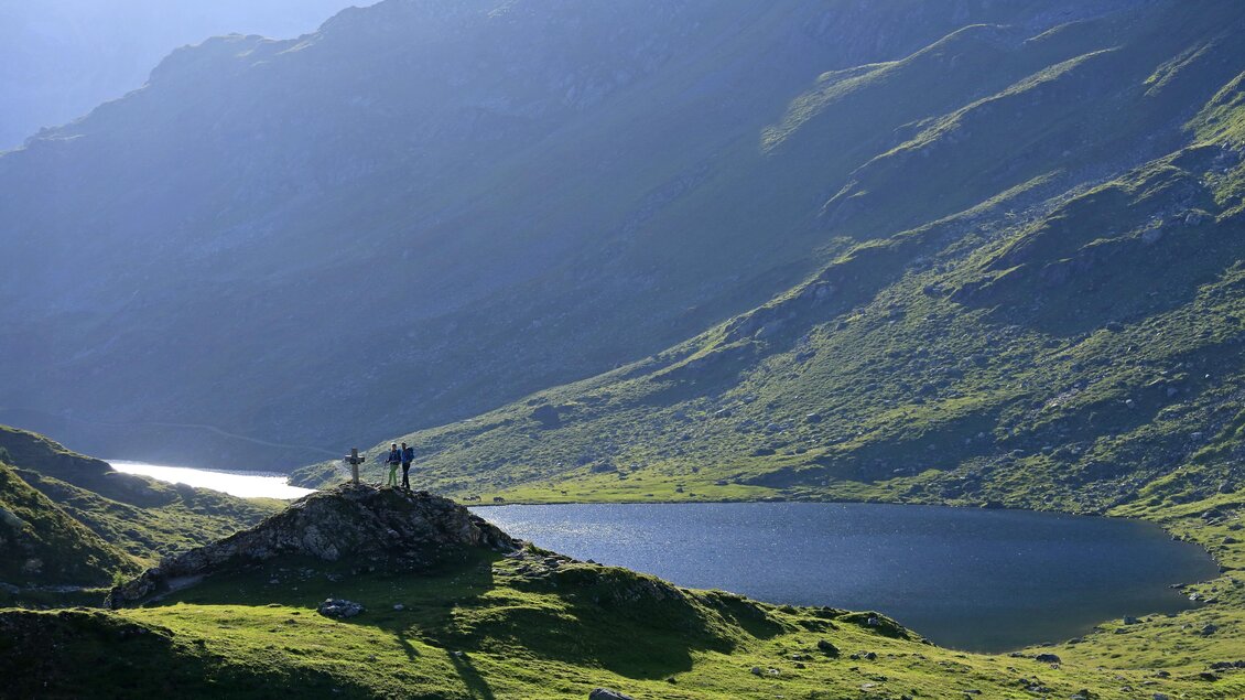 Eine malerische Berglandschaft mit einem ruhigen See und sanften Hügeln. Einige Menschen stehen auf einem Felsen und genießen die Aussicht. | © Herbert Raffalt