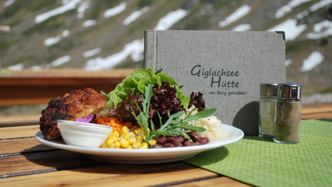 Ein Teller mit einer Vielzahl von frischem Gemüse, Salat und einer herzhaften Beilage auf einem Tisch. Im Hintergrund steht ein Buch mit dem Titel „Gulachsee Hütte“. | © Gigalchseehütte