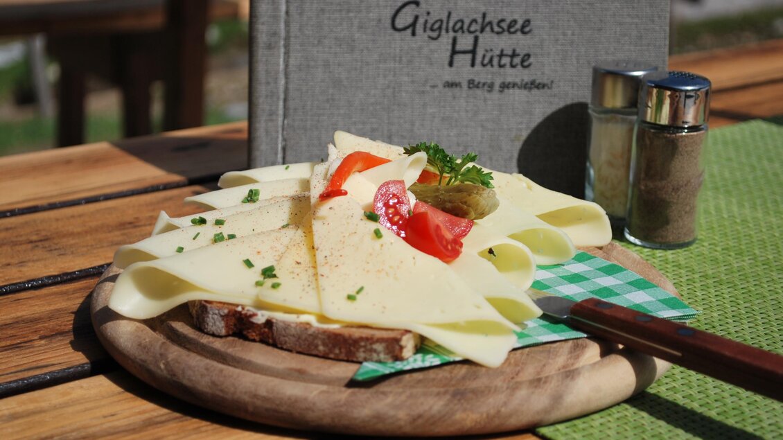 Ein Teller mit frischem Brot, belegt mit Käse, Tomaten und Kräutern. Im Hintergrund steht eine graue Speisekarte der Giglachensee Hütte. | © Gigalchseehütte