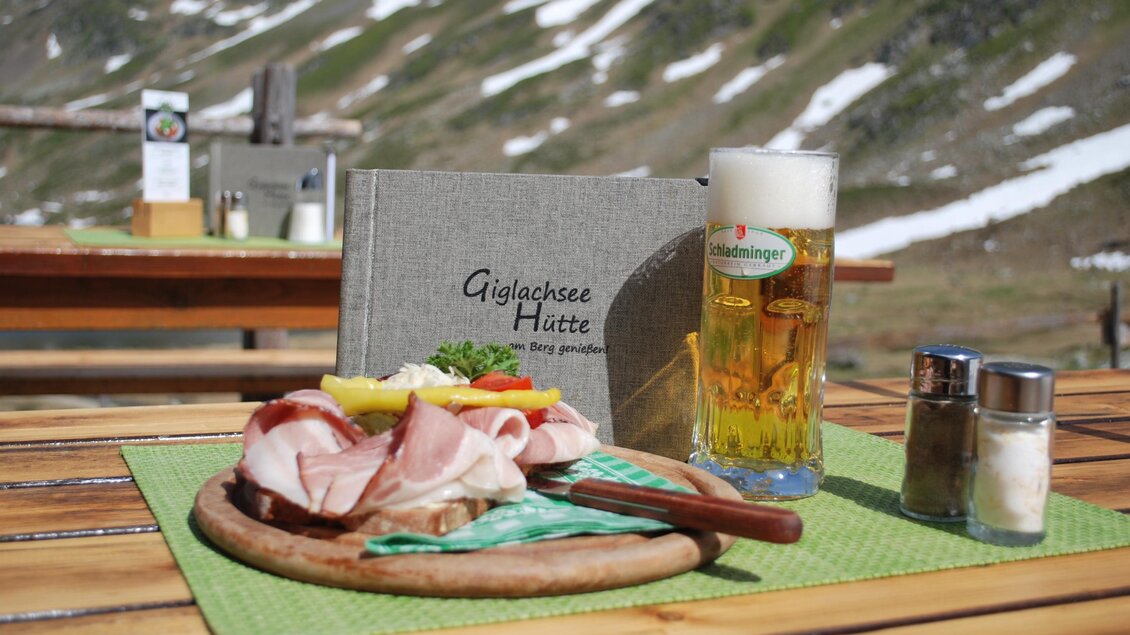 Ein Teller mit Wurst und Käse, garniert mit frischem Gemüse, steht auf einem Holztisch. Dazu gibt es ein Glas Bier und eine Speisekarte inmitten einer bergigen Landschaft. | © Gigalchseehütte