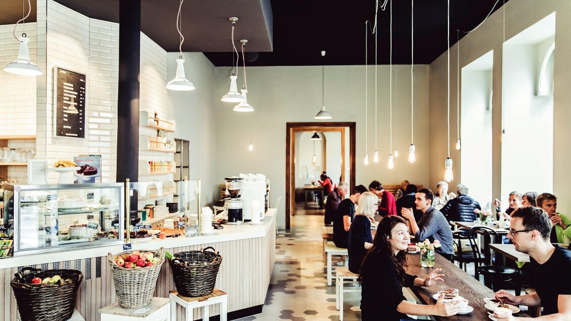 Ein modernes Café mit einem hellen Innenraum. Gäste sitzen an Tischen und genießen Getränke und Speisen. | © Ginko - Lupi Spuma