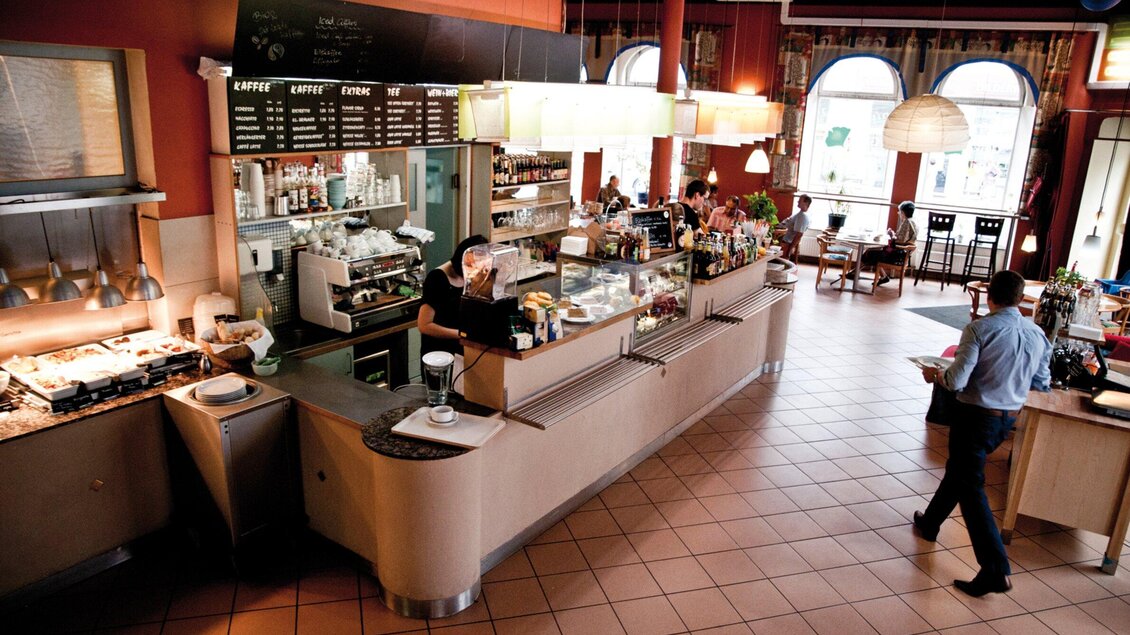 Ein modernes Café mit einer langen Theke und verschiedenen Sitzbereichen. Angestellte bedienen Gäste in einer einladenden Atmosphäre. | © Ginko - Udo Stelzer