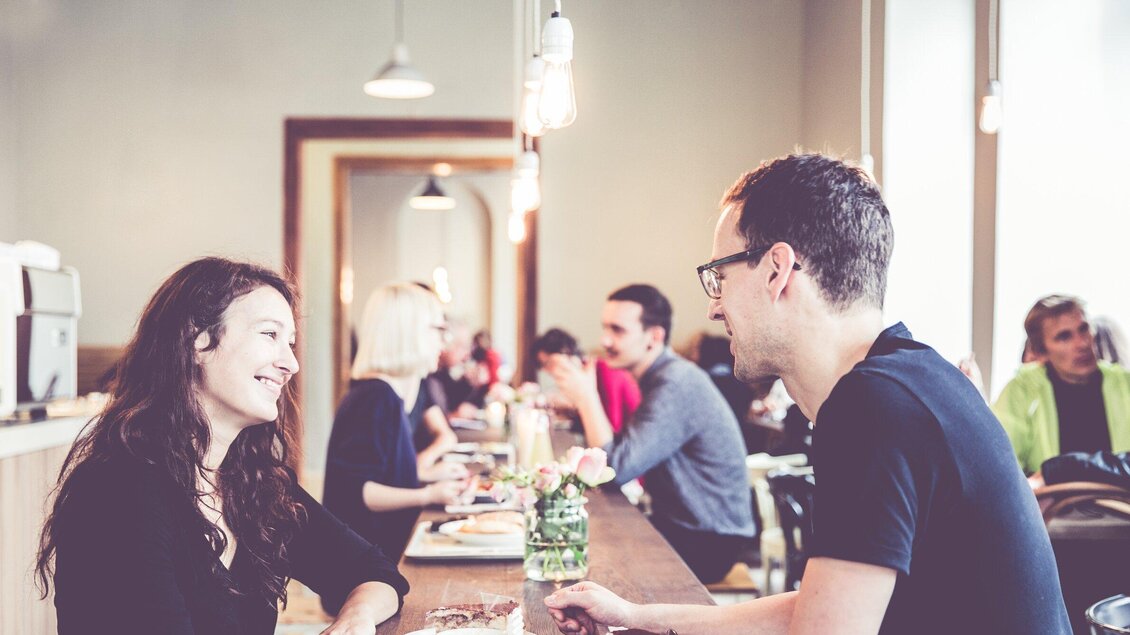Ein modernes Café mit mehreren Gästen, die entspannt essen und trinken. Ein Paar führt ein fröhliches Gespräch an einem Tisch. | © Ginko - Lupi Spuma