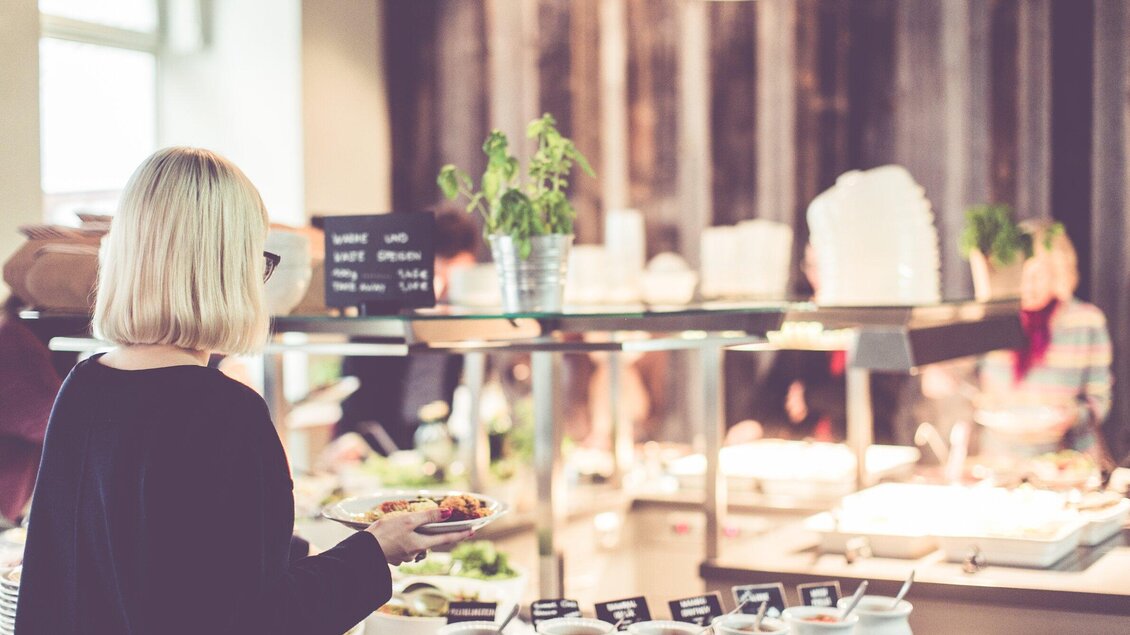 Eine Person mit blonden Haaren steht an einem Buffet und hält einen Teller mit Essen. Im Hintergrund sind verschiedene Gerichte und andere Gäste sichtbar. | © Ginko - Lupi Spuma
