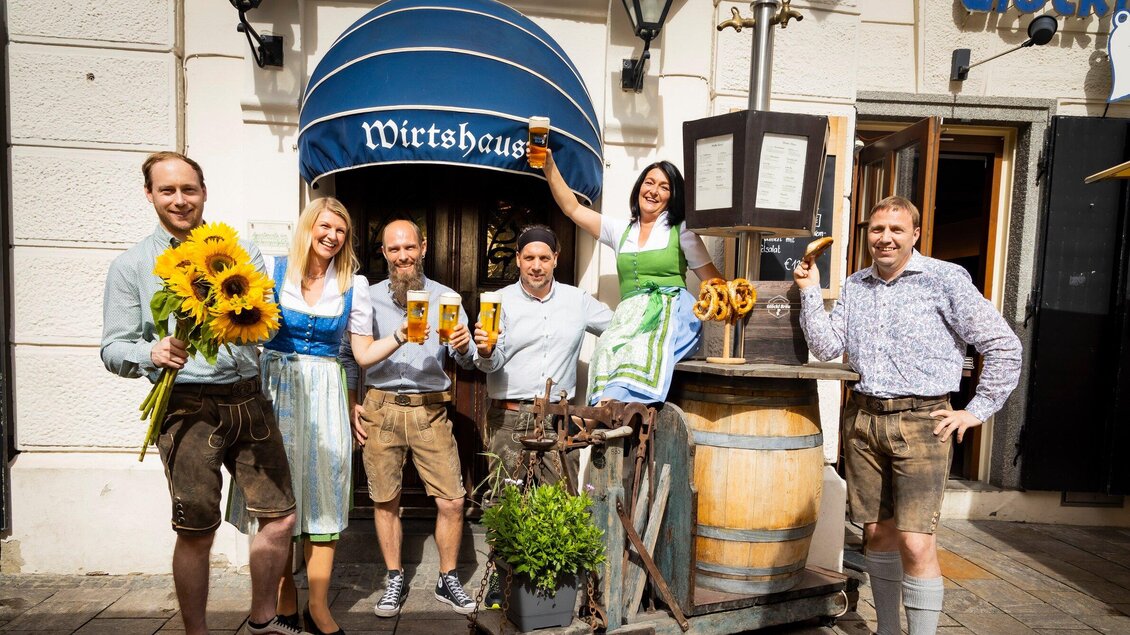 Eine fröhliche Gruppe von Menschen steht vor einem Wirtshaus und hält Getränke und Sonnenblumen in den Händen. Sie tragen traditionelle bayerische Trachten. | © Glöckl Bräu