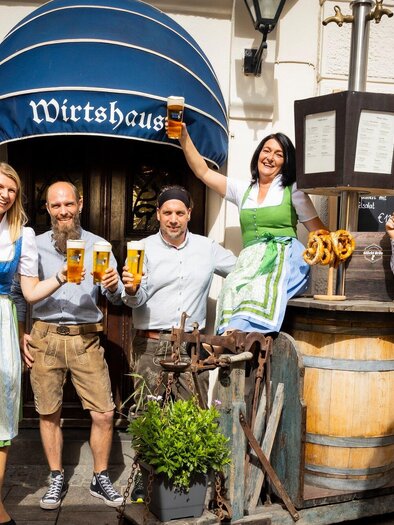 Eine fröhliche Gruppe von Menschen steht vor einem Wirtshaus und hält Getränke und Sonnenblumen in den Händen. Sie tragen traditionelle bayerische Trachten. | © Glöckl Bräu