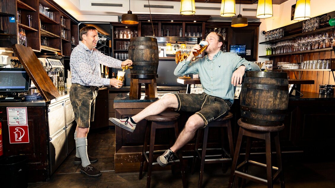 Zwei Männer in traditioneller bayerischer Tracht genießen Bier in einer gemütlichen Bar. Im Hintergrund sind Holzfässer und eine einladende Atmosphäre zu sehen. | © Glöckl Bräu