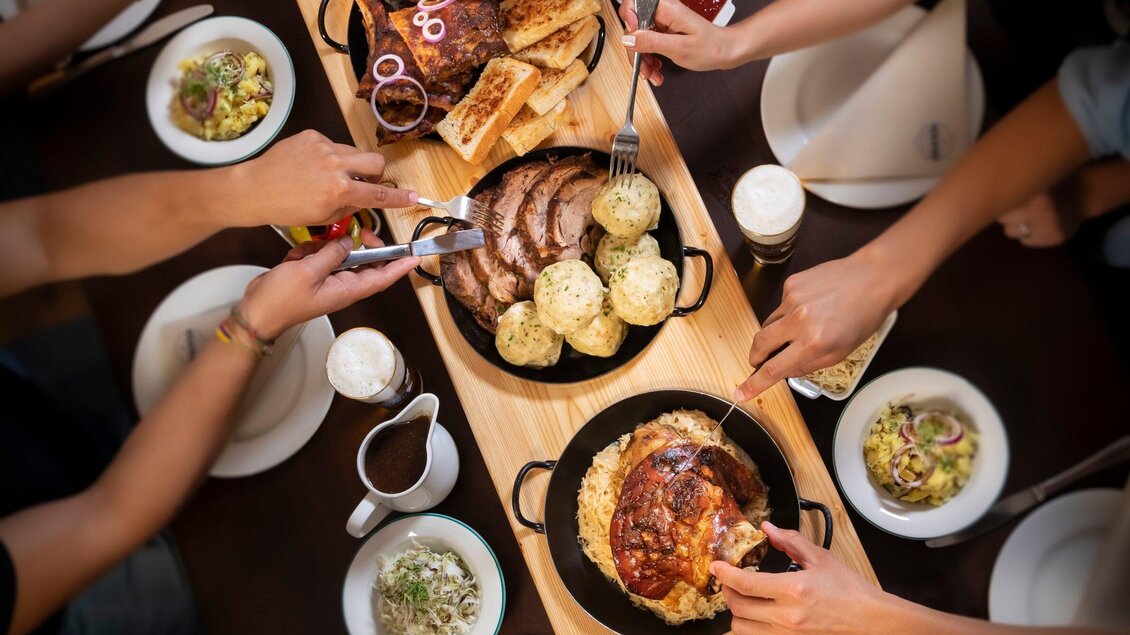Eine schöne gedeckte Tafel mit verschiedenen Fleischgerichten und Beilagen. Mehrere Hände greifen nach dem Essen, was eine gesellige Atmosphäre schafft. | © Glöckl Bräu