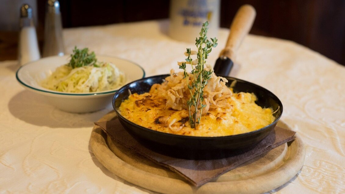 Ein herzhaftes Gericht in einer schwarzen Pfanne mit Käse und Zwiebeln, garniert mit frischen Kräutern. Nebenbei liegt eine Schüssel mit Salat. | © Glöckl Bräu