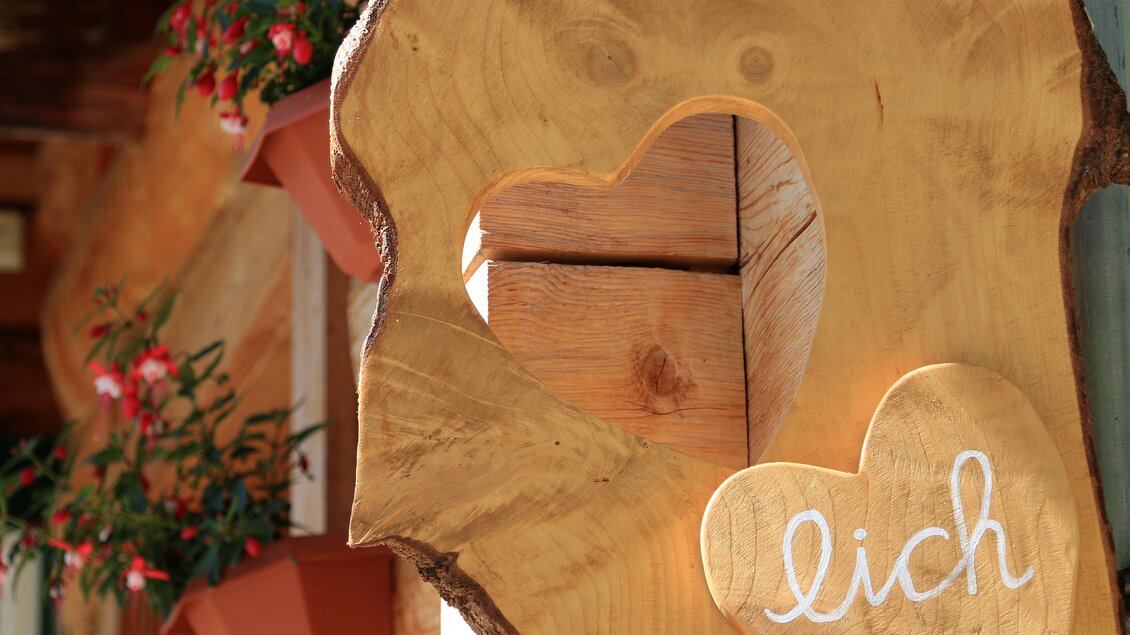 Ein Holzschild mit einem Herz-Ausschnitt und dem Schriftzug „lich“. Im Hintergrund sind blühende Pflanzen in Töpfen zu sehen. | © Glösalm