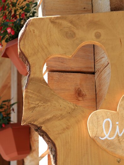 A wooden sign with a heart-shaped cutout and the inscription "lich." In the background, blooming plants in pots can be seen. | © Glösalm