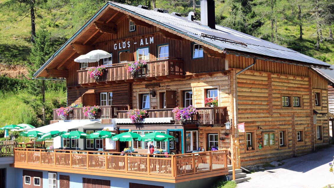 Ein gemütliches Holzhaus mit Balkonen, das bunt mit Blumen geschmückt ist. Auf der Terrasse stehen grüne Schirme und Stühle für Gäste. | © Glösalm