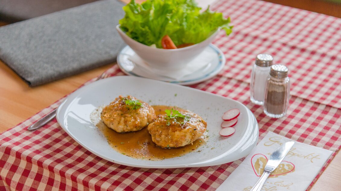 Ein Teller mit zwei gebratenen Fischfilets in Sauce, garniert mit Kräutern. Im Hintergrund liegt ein Schüssel mit frischem Salat und eine Tischdecke im karierten Muster. | © Glösalm