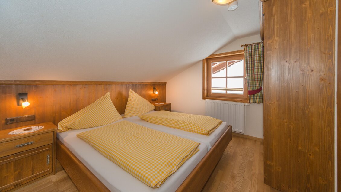Ein gemütliches Schlafzimmer mit einem Doppelbett und Holzvertäfelung. Das Fenster lässt Tageslicht herein und sorgt für eine freundliche Atmosphäre. | © Glösalm