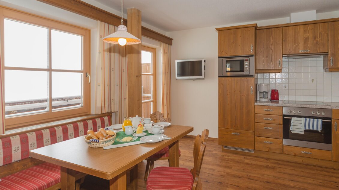 Eine gemütliche Küche mit einem Esstisch und Holzmöbeln. Im Hintergrund sind Fenster mit Blick nach draußen und eine moderne Küchenausstattung sichtbar. | © Glösalm