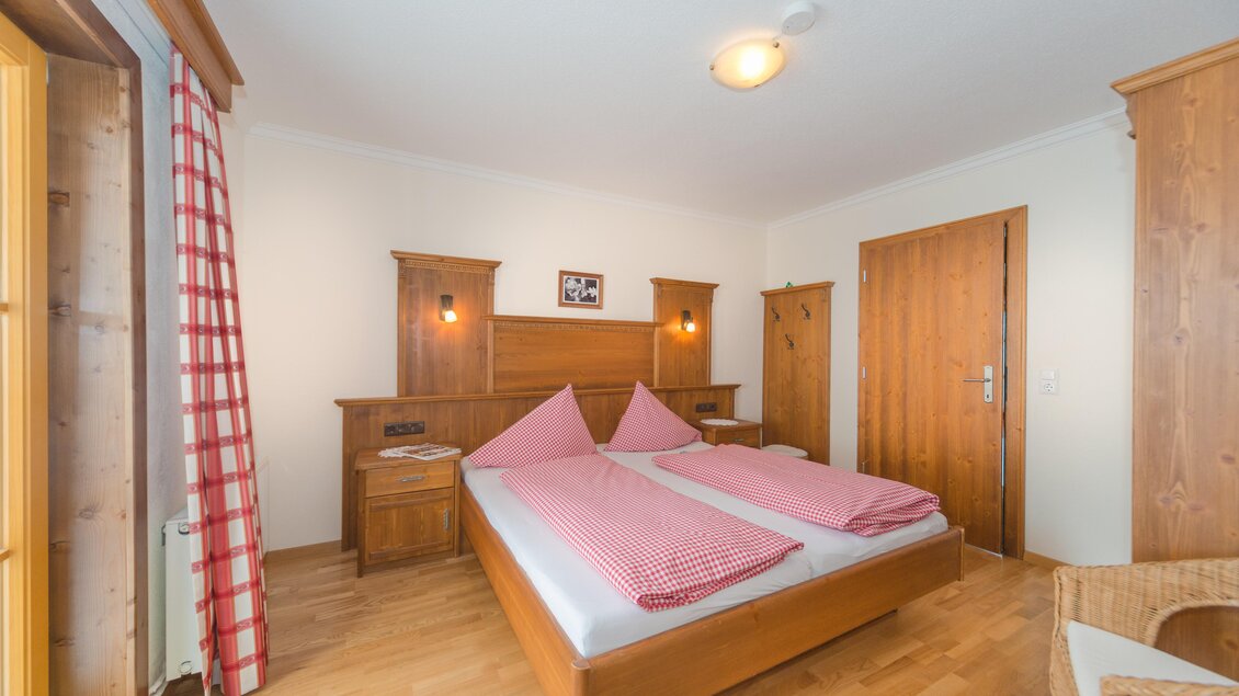 Ein gemütliches Schlafzimmer mit zwei Einzelbetten und Holzmöbeln. Die Wände sind hell und es gibt Fenster mit roten Vorhängen. | © Glösalm
