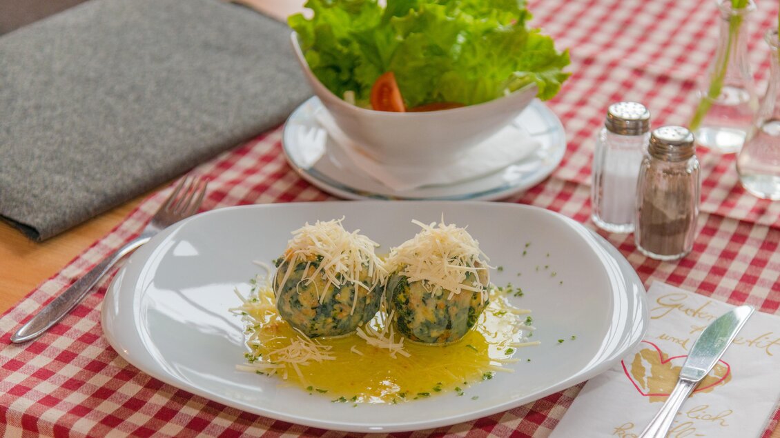 Ein Teller mit zwei Spinatknödeln, garniert mit Parmesan und umgeben von einer feinen Ölsauce. Im Hintergrund ist ein Salat in einer Schüssel zu sehen. | © Glösalm