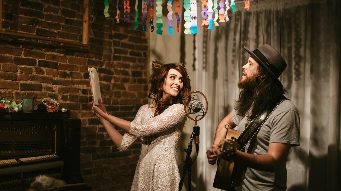 Ein Paar Musiker in einem gemütlichen Raum mit Backsteinmauer. Die Frau trägt ein weißes Kleid und hält ein Mikrofon, während der Mann Gitarre spielt. | © Glam Feldbach