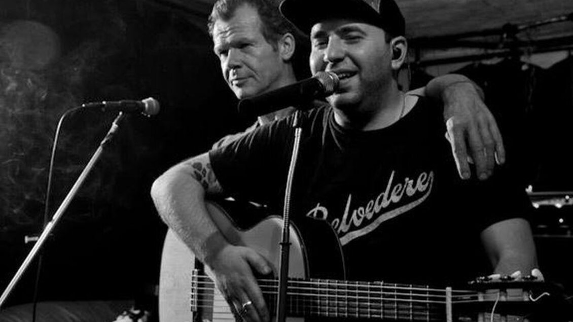 Zwei Musiker stehen auf der Bühne und lachen. Der eine spielt Gitarre, während der andere ihn freundlich anschaut. | © Glam Feldbach