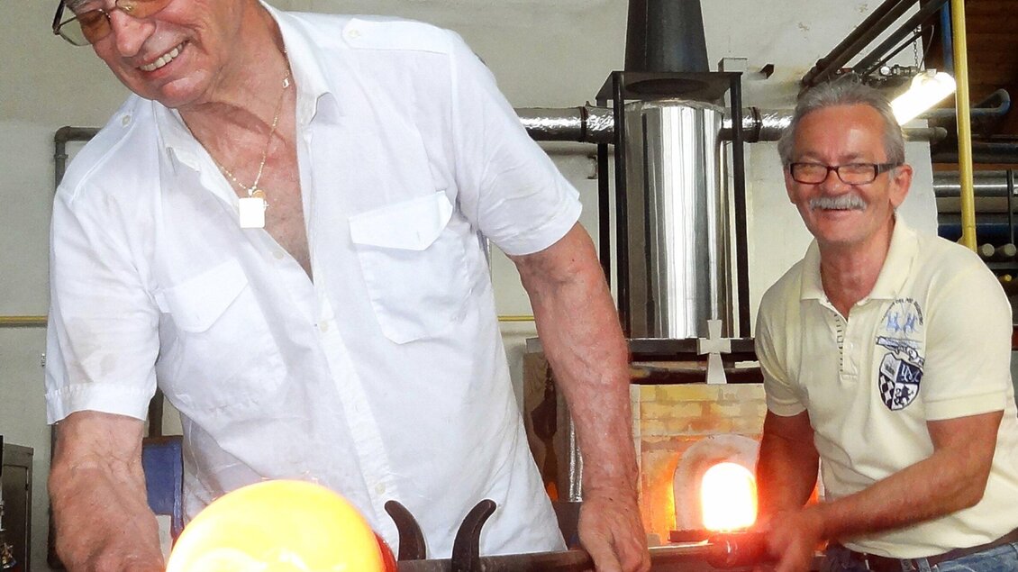 Zwei Männer arbeiten gemeinsam in einer Glasbläserwerkstatt. Sie formen glühendes Glas und lächeln dabei. | © GlasMuseumBärnbach