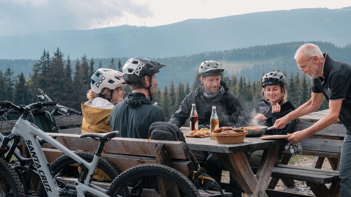 Eine Gruppe von Radfahrern sitzt an einem Tisch im Freien und genießt eine Mahlzeit in der Natur. Im Hintergrund sind eine bewaldete Landschaft und Berge zu sehen. | © Verein Tourismusentwicklung Steirischer Wechsel