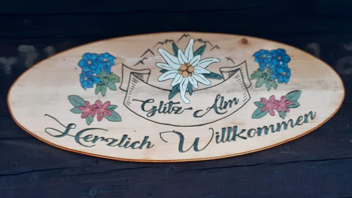 Ein handgefertigtes Holzschild mit der Aufschrift "Glitz-Alm" und "Herzlich Willkommen". Umgeben von bunten Blumen und Bergmotiven. | © Glitzhütte