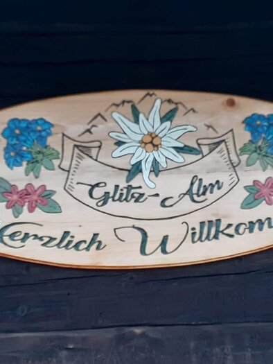 A handcrafted wooden sign with the inscription "Glitz-Alm" and "Welcome". Surrounded by colorful flowers and mountain motifs. | © Glitzhütte