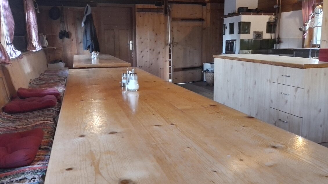 Eine gemütliche Holzküche mit einem langen Tisch und roten Kissen. Die Wände sind aus Holz und es gibt eine offene Küche im Hintergrund.