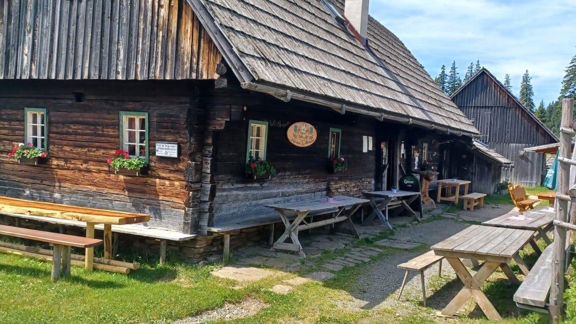 Ein rustikales Holzhaus mit Blumenkästen und mehreren Sitzgelegenheiten im Freien. Die Umgebung ist grün und bietet einen sonnigen, einladenden Eindruck. | © Fam. Lipp