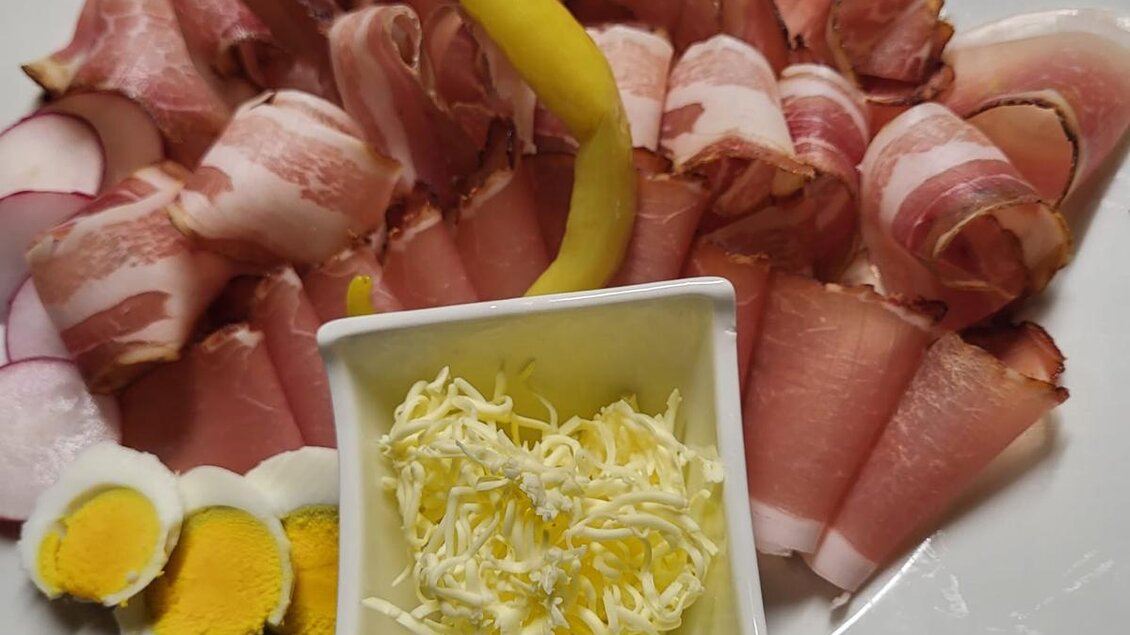 Ein Teller mit verschiedenen Wurstspezialitäten, darunter Fleisch und Speck, garniert mit Gurken, Tomaten und Radieschen. In der Mitte befindet sich eine Schale mit geriebenem Käse in Herzform. | © Fam. Lipp