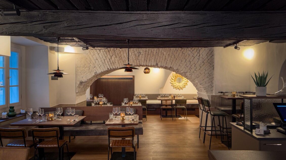 Innenansicht eines eleganten Restaurants mit Holzbalkendecke, architektonischen Bögen und modernen Tischen, die mit Weingläsern und Besteck gedeckt sind. | © Glockenspielkeller
