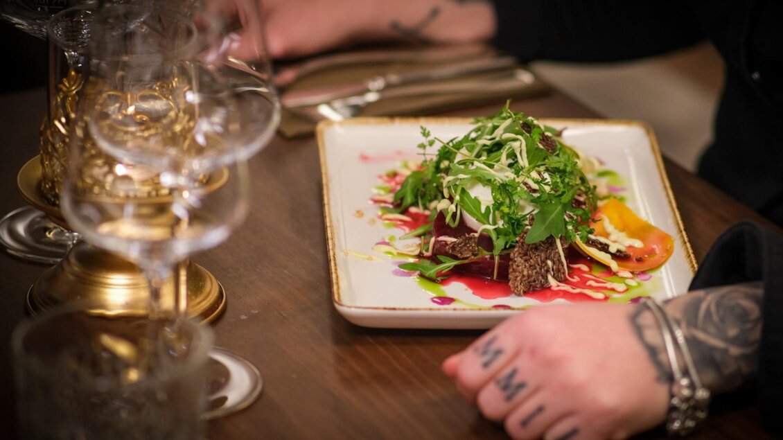 Ein kunstvoll angerichtetes Gericht mit frischen Kräutern und einer bunten Soßendekoration, begleitet von edlen Weingläsern und einer tätowierten Hand. | © Glockenspielkeller