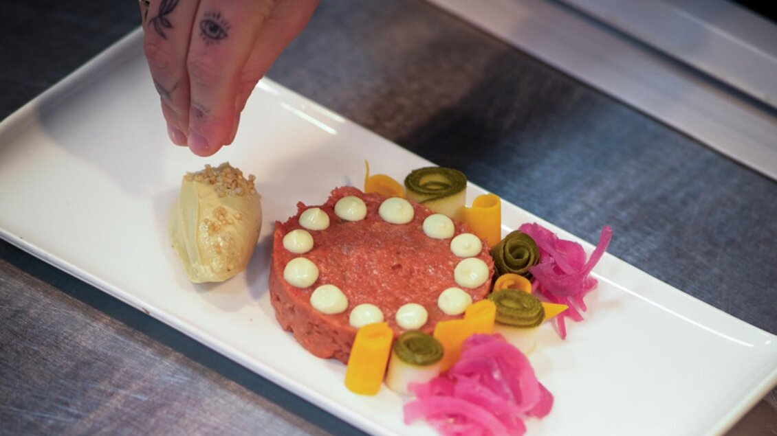 Ein kunstvoll angerichteter Teller mit rohem Fleisch, verziert mit buntem Gemüse, Kräutern und Saucen, während eine Hand die letzte Feinheit hinzufügt. | © Glockenspielkeller