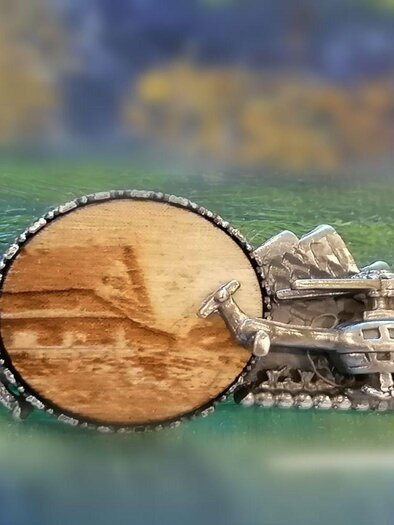 Eine dekorative Auszeichnung mit einem Holzfeld und einem silbernen Hubschrauber. Im Hintergrund sind stilisierte Berge und Tannen dargestellt. | © Anita Seebacher