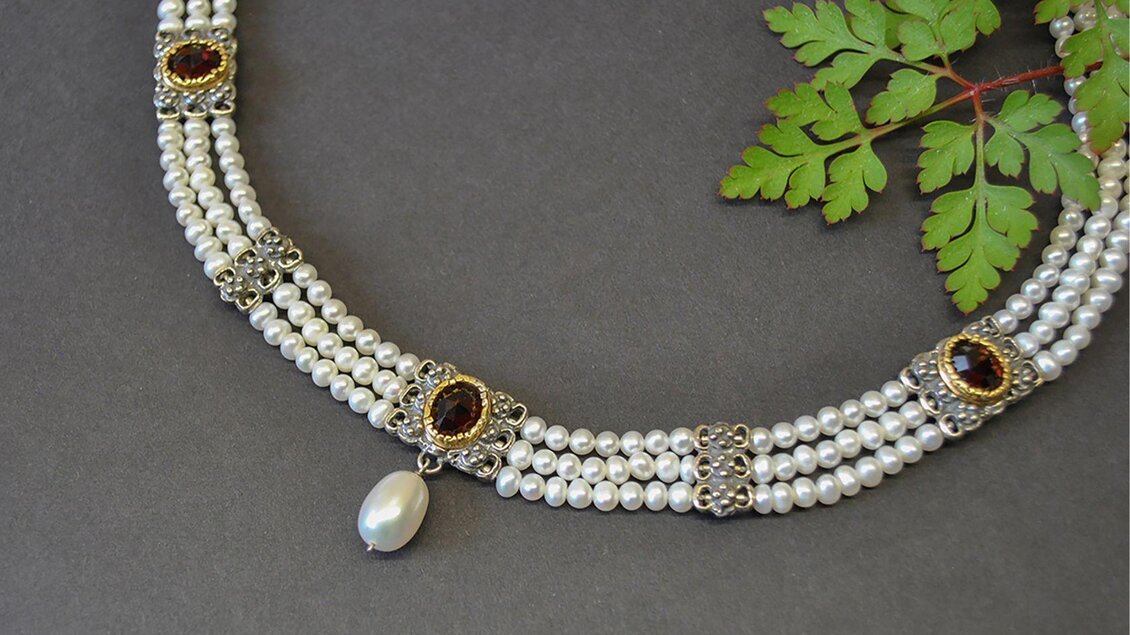 Eine elegante Halskette aus weißen Perlen mit geschmackvollen Verzierungen und einem Anhänger. Sie liegt neben einem grünen Blatt auf einem dunklen Hintergrund. | © Goldschmiede Steiner
