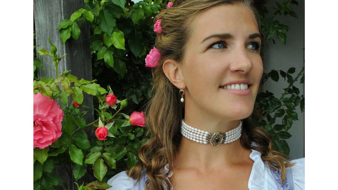 Eine junge Frau mit lockigem Haar und rosa Blumen im Haar lächelt freundlich. Sie trägt eine Perlenkette und steht vor einem Hintergrund von grünen Pflanzen und Rosen. | © Goldschmiede Steiner