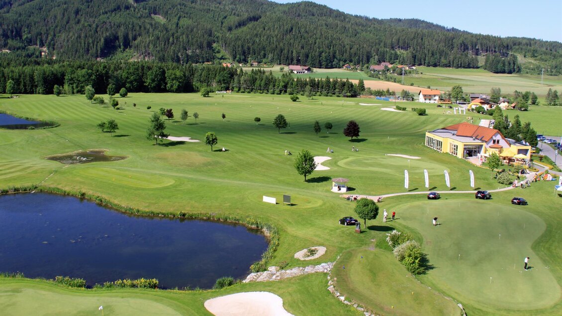 Eine weitläufige Golfanlage mit gepflegten Rasenflächen und einem Teich. Im Hintergrund ist ein Gebäude und Wald zu sehen. | © Golf Club Murtal