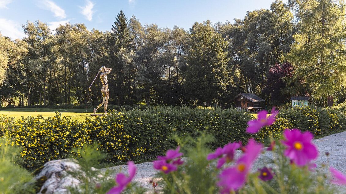 Eine Statue auf einer grünen Wiese, umgeben von Bäumen und bunten Blumen. Der Himmel ist blau und der Ort wirkt einladend und friedlich. | © Thomas Sattler