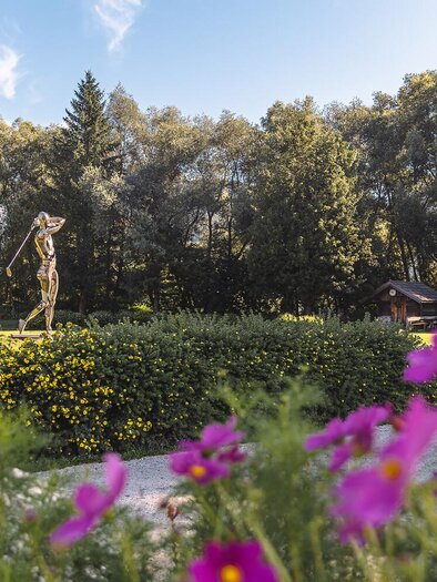 A statue on a green meadow, surrounded by trees and colorful flowers. The sky is blue and the place feels inviting and peaceful. | © Thomas Sattler