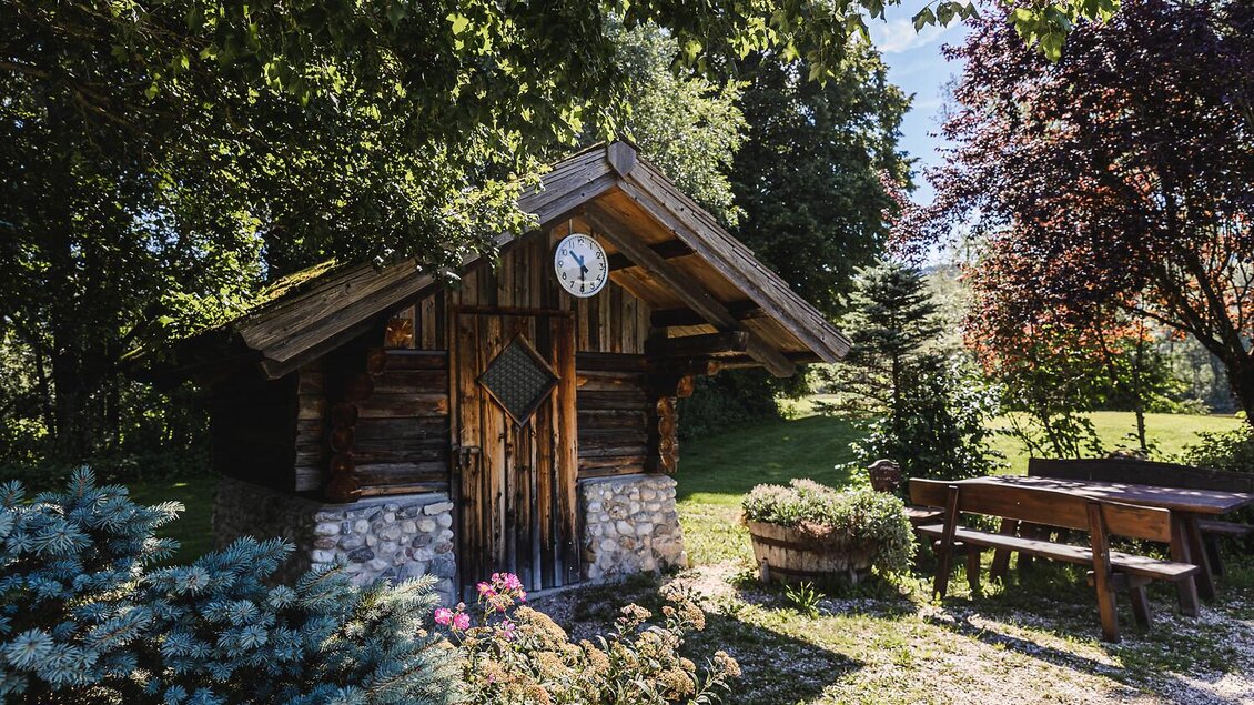 Ein rustikales Holzhaus in einer grünen Umgebung. Im Vordergrund sind Blumen und ein Tisch mit Bänken zu sehen. | © Thomas Sattler