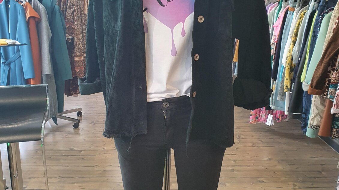 Ein modisches Outfit bestehend aus einer schwarzen Jacke, einem T-Shirt mit Herzmotiv und einer schwarzen Hose. Der Hintergrund zeigt eine Kleiderboutique mit bunten Kleidungsstücken. | © Golf-Fashion-Lifestyle Judith Schraberger