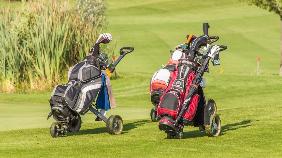 Zwei Golfcaddies stehen auf einer grünen Golfbahn. Im Hintergrund sind hohe Gräser und ein klarer, blauer Himmel zu sehen. | © Ingrid Jansky