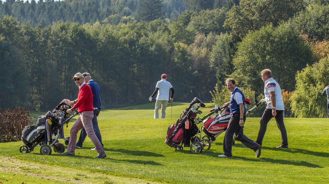 Eine Gruppe von Golfern geht mit ihren Golfbags über ein grünes Golfplatzgelände. Im Hintergrund sind Bäume und ein klarer Himmel zu sehen. | © Ingrid Jansky