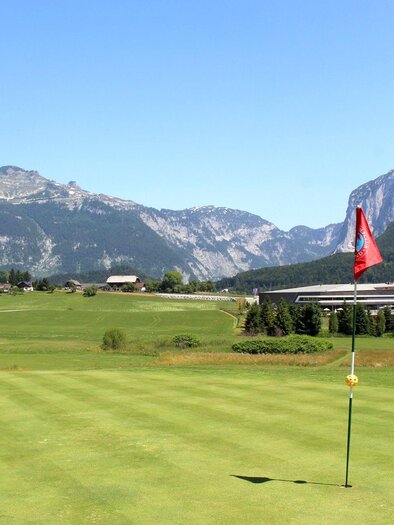 A picturesque golf course with well-maintained greens and a flagpole. In the background, majestic mountains rise under a clear blue sky. | © TVB Ausseerland - Salzkammergut_Viola Lechner