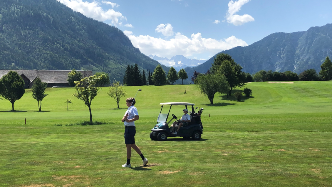 Ein schöner Golfplatz mit grünen Wiesen und Bergen im Hintergrund. Ein Spieler steht bereit, während ein Golfcart vorbeifährt. | © Astrid Wichert
