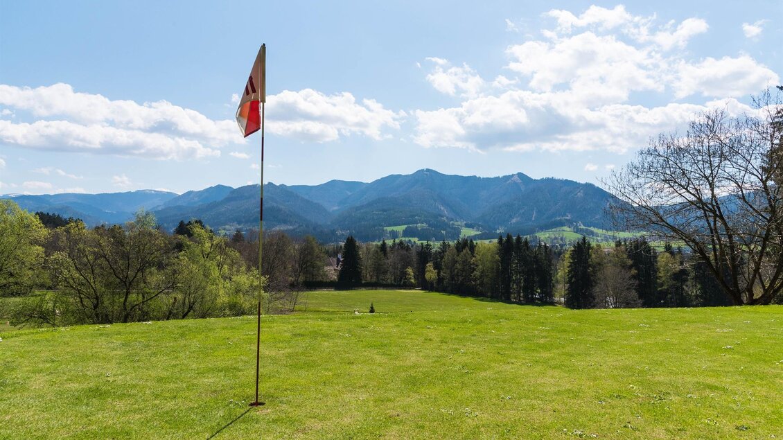 Eine weitläufige grüne Wiese mit einer Fahne im Vordergrund und Bergen im Hintergrund. Der Himmel ist klar und sonnig. | © Golfclub Schloss Feistritz