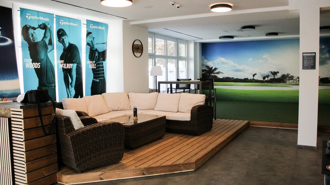 Ein modern eingerichteter Raum mit einem gemütlichen Sofa und einem Holzboden. An der Wand sind Bilder von Golffiguren und ein grüner Golfplatz abgebildet. | © Golfstudio ML
